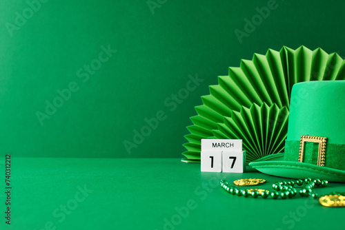 St. Patrick's Day revelry: a touch of emerald charm. Side view of green hat, calendar set to March 17th, green paper fan decoration, gold coins on green background with space for festive announcements
