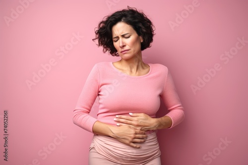  Photography a middle-aged woman holding her belly with a worried expression, against a solid pastel pink background, portraying the discomfort of her stomach pain