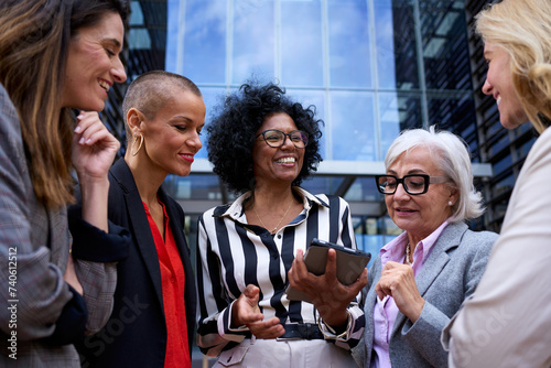 Team diverse ages and race business women conversing cheerful standing outside office building. Professionals successful positive mature females talking about commerce looking and using tablet in hand