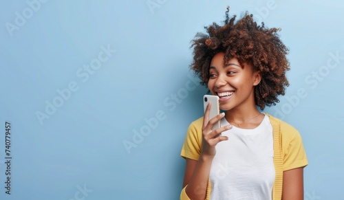 Half length shot of Afro woman holds mobile phone, enjoys nice talk online in social networks, reads funny article in internet, uses cellular isolated over blue background.