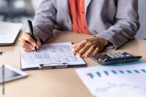 Close up of unrecognizable black woman in formal outfit filling up tables on clipboard next to electronic calculator. Responsible administrator entering data by hand with pen on wooden desk