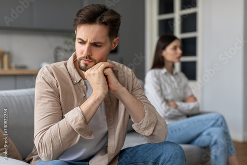 Unhappy young man sitting on sofa separate from discontented wife on background