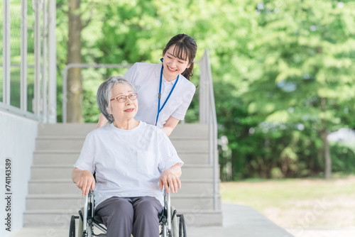 公園を散歩する車椅子に乗った高齢者と介護士（ヘルパー） 