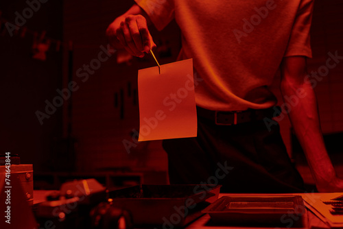 cropped shot of photographer holding tweezers with photo paper in a darkroom with red light