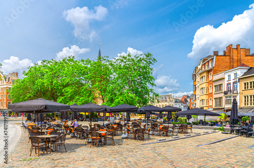 Street restaurant with tables, chairs and umbrellas, green trees and old buildings on Grote Markt square in Kortrijk city historical centre in sunny summer day, West Flanders province, Belgium