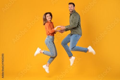 Jubilant man and woman holding hands and jumping in excitement, capturing joyful moment in full-length shot against vibrant yellow background