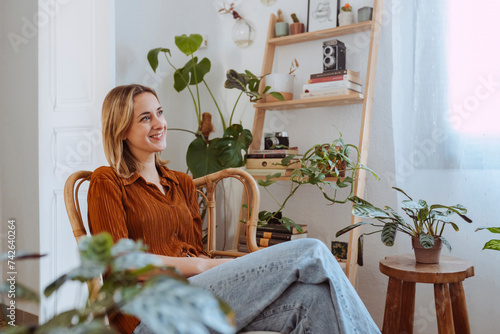 Portrait of a young cheerful woman sitting on the chair in her living room early in the morning and looking at the window. Copy space, interior background.