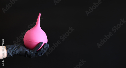 A hand in a black latex glove holds a large pink enema on a dark background. Medical device. Pear-shaped enema, body cleansing concept