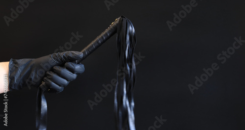 A hand in a black latex glove holds a whip against a dark background. Leather whip for spanking isolated on black. Sex toy for intimacy. Sexual slavery