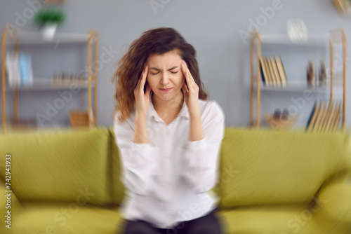 Woman feels dizzy and sits down on the couch. Young woman sitting on the sofa at home, feeling pain and a spinning sensation in her head. Concept of headache, vertigo, health problem, brain tumor