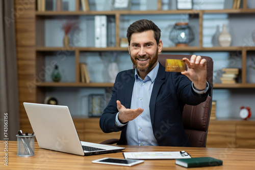 Portrait of a smiling young successful businessman, banker, financial director sitting at an office desk in a suit, holding and showing a golden plastic credit card to the camera