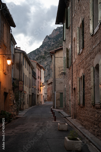 Vertical photograph of the picturesque French winding alley of Villefranche de Conflent at dusk. There is a street lamp with warm light