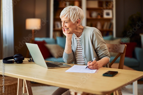 A cheerful senior adult woman working from home using laptop and writing reports
