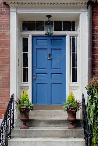 A vibrant blue front door features sidelight windows on each side and a transom window above. Two potted plants outside the door in New England.
