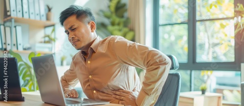 Asian businessman having sick back pain after extra time working with laptop in office. AI generated
