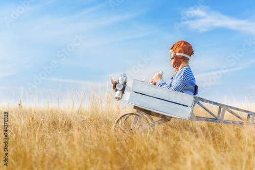 Little boy is playing the plane. Happy and funy child imagines himself an aircraft pilot and plays in a aviator costume in an open-air field against a blue sky on a summer sunny day. 