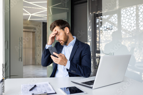 Shocked young man businessman sitting in the office on a chair and holding his head with his hand, holding the phone, upset by the news he received