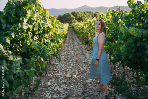 A girl in a blue sundress stands on a vine alley in a vineyard. Mountains in the background, sunset. Vineyards at the winery