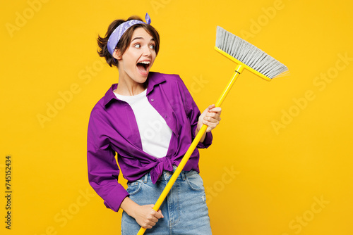 Young shocked excited happy fun woman she wear purple shirt casual clothes do housework tidy up hold in hand broom look aside isolated on plain yellow background studio portrait. Housekeeping concept