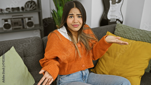 Puzzled young woman with outstretched hand, sitting in a modern living room, looking unsure or questioning.