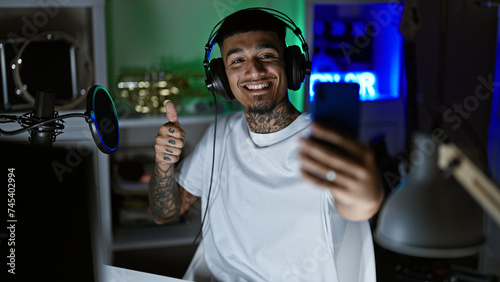 Smiling, tattooed young latin man, a music studio maestro, snapping a vibrant selfie with his smartphone amidst the recording session, donning his headphones. a solo night cap in his artistic haven.