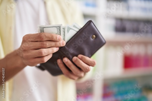 Young hispanic man customer holding dollars of wallet at pharmacy