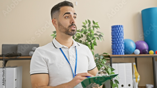 A young hispanic man with a beard, wearing a lanyard, holds a clipboard in an indoor rehabilitation clinic setting.