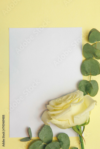 Kartka papieru na żółtym tle, róża i eukaliptus położona na białej kartce. Miejsce na tekst.