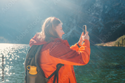 Rear shot of traveler woman influencer in sportswear with backpack standing on the rocky shore and making selfie on smart phone. Taking photo of green hills and mountains on Morskie Oko lake. Hiking