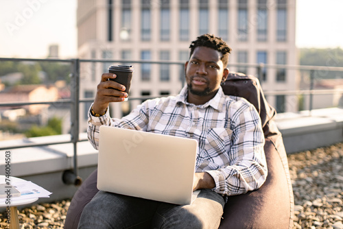 Adult businessman in casual wear carrying out professional activities in open air. Portrait of African male drinking coffee while placing laptop on knees sitting on bean bag chair on roof terrace.