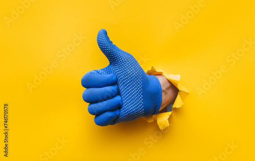 A right man's hand in a blue fabric work glove shows a thumb up (like). Torn hole in yellow paper. Good job, fun mood and approval concept. Copy space.