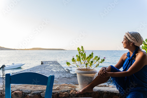 Beautiful woman with blue sarong and Greek outfit on her head relaxingly enjoying the spectacular view of the sea to the south of the greek island of Crete in summer from a Greek tavern