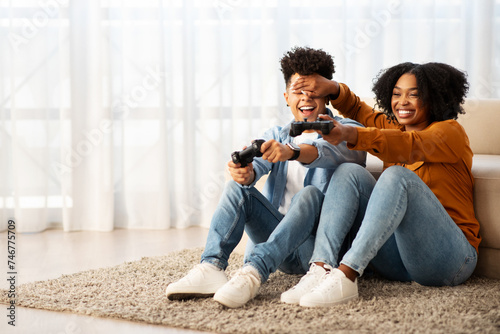 Joyful African American couple playing video games on the floor, with the woman playfully covering the man's eyes