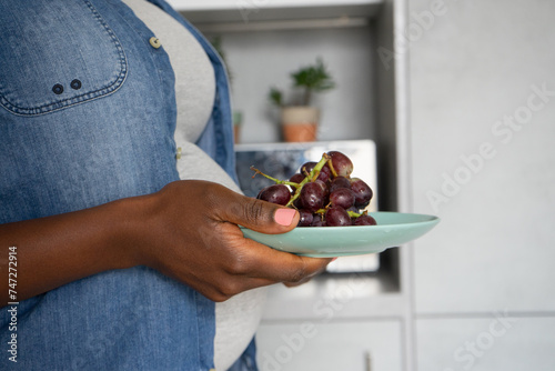 Mid section of pregnant woman holding plate with red grapes in kitchen