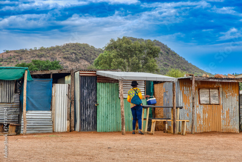 township informal settlement in africa near a hill, woman caring a cooler box in front of the corrugated iron shacks