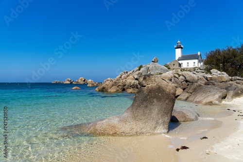 Sur une plage de sable, plusieurs rochers, dont un en forme de pied, émergent des eaux cristallines. Le phare de Pontusval se dresse sur une pointe rocheuse en Bretagne.