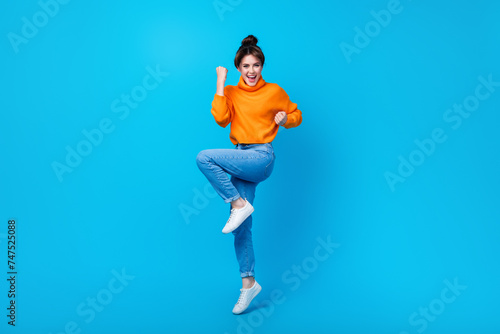 Full length photo of cute lucky woman dressed orange sweater rising fists jumping high isolated blue color background