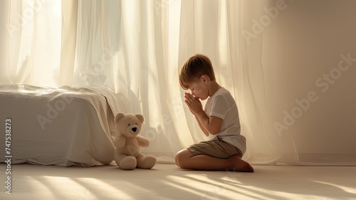 a three-year-old boy kneeling in prayer beside his bed in his bedroom, head bowed, with a teddy bear nearby, against a backdrop of soft white pastel colors.