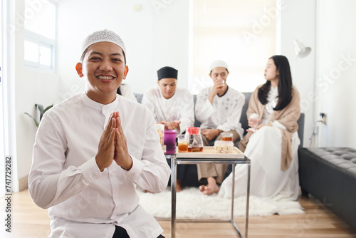 Friendly Asian muslim man wear skullcap showing greeting or welcome gesture during Eid mubarak celebration and his friends gather on the background