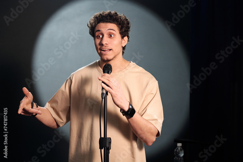 Young comedian of stand up club standing on stage in front of audience and pronouncing monologue in microphone during performance