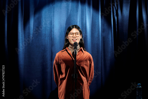 Young Chinese female comedian in brown shirt standing on stage with blue curtains and speaking in microphone during monologue