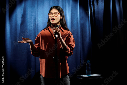 Young comedian in eyeglasses and brown shirt standing on stage against blue curtains and speaking in microphone during performance