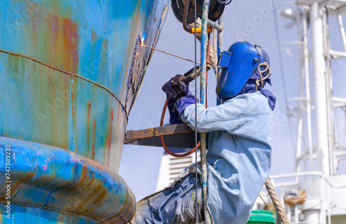 Welder rappelling with ladder to welding metal hull surface of the old fishing boat during maintenance and improvement work in shipyard area