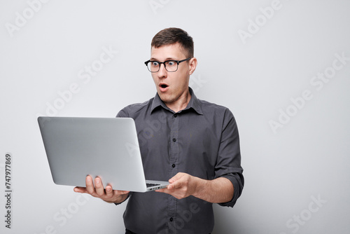 Surprised young man in glasses holding laptop with open mouth expression isolated on gray background
