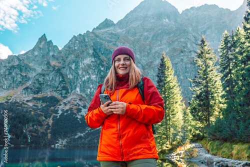 Shot of happy woman looking at the phone with backpack relaxing outdoor with rocky on Morskie Oko lake, hiking mountains. Travel and hobby concept. 