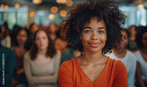 A confident black woman with curly hair stands proudly at the forefront of a lively crowd, sporting an orange top. Her empowering presence shines as she leads with charisma and determination, inspirin