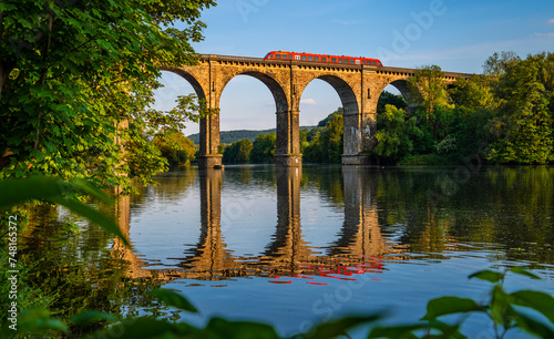 Panoramic view of railway viaduct with train crossing Ruhr river in Herdecke Germany. Historic brick construction with tall arches on the branch line from Hagen to Dortmund reflected by water surface.