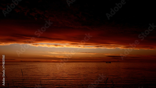 Fairytale breathtaking sunset above rippling river with a floating boat. Clip. Dark red and orange sky, heavy clouds above water.