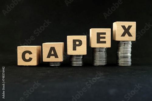 Increase in capex or capital expenditure concept. Wooden blocks with increasing stack of coins in black background.