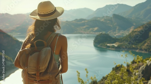 woman with a hat and backpack looking at the mountains and lake from the top of a mountain in the sun light, with a view of the mountains 